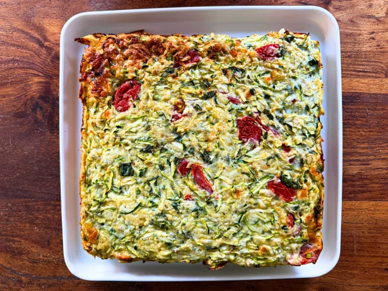 Overhead view of a classic baked zucchini slice in a white rectangular dish, made with grated zucchini, fresh basil, melted cheese and juicy cherry tomatoes, lightly golden on top and around the edges, set on a rustic wooden surface.