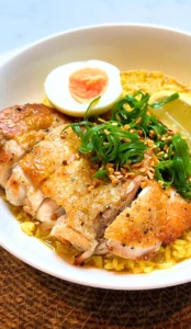 Chicken rice dish, with golden-skinned sliced roasted chicken, a soft-boiled egg half, and aromatic yellow rice, garnishes with finely shredded green onions, crispy fried garlic, and a fresh lemon wedge in a white ceramic bowl, set on a rustic woven mat under natural lighting.